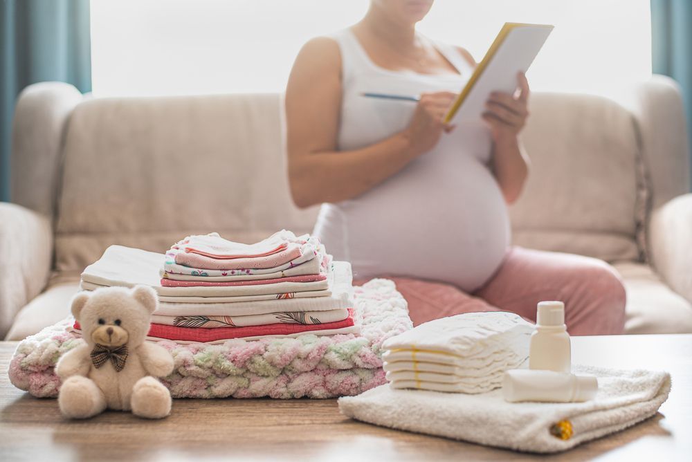 A pregnant woman is sitting on a couch with a teddy bear and diapers.