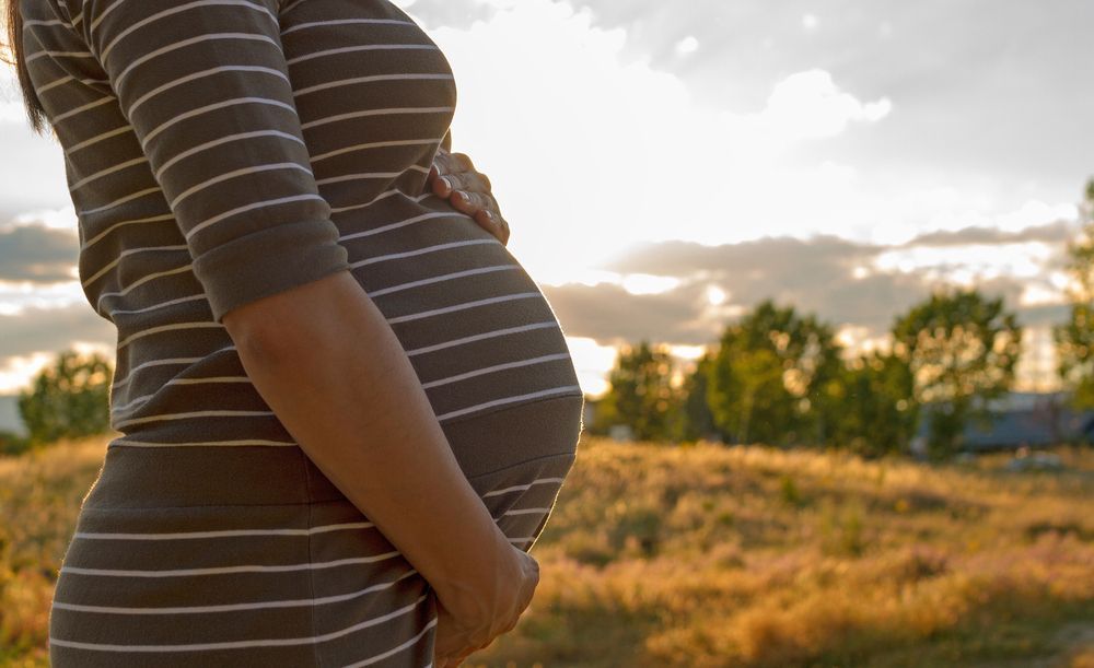 A pregnant woman in a striped dress is standing in a field holding her belly.