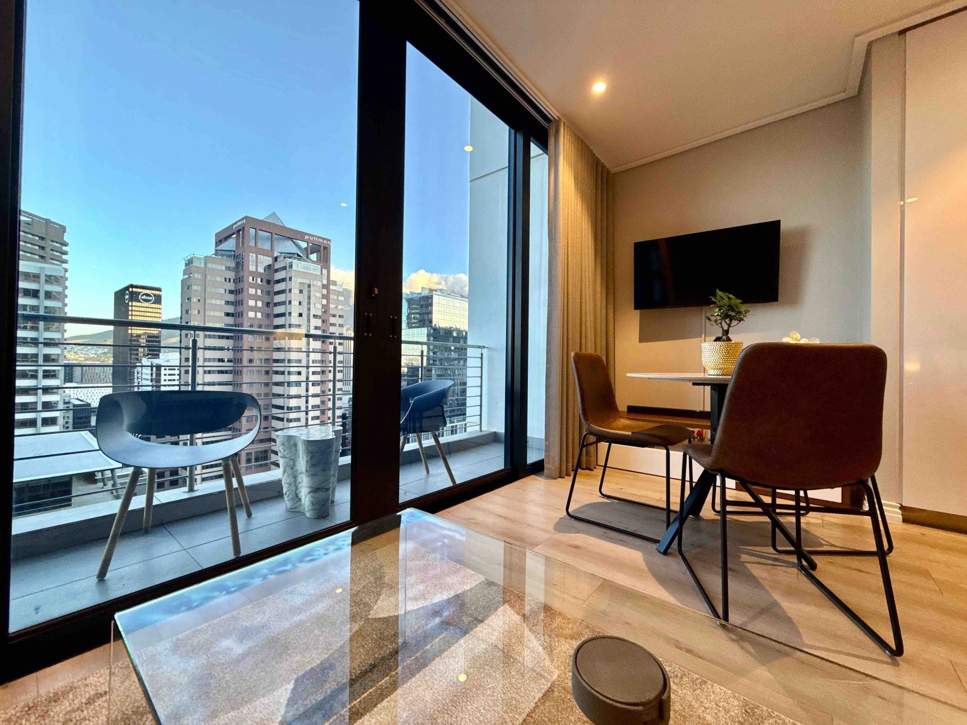 Modern apartment balcony and living room with city view, glass doors, chair, table, and TV bench.