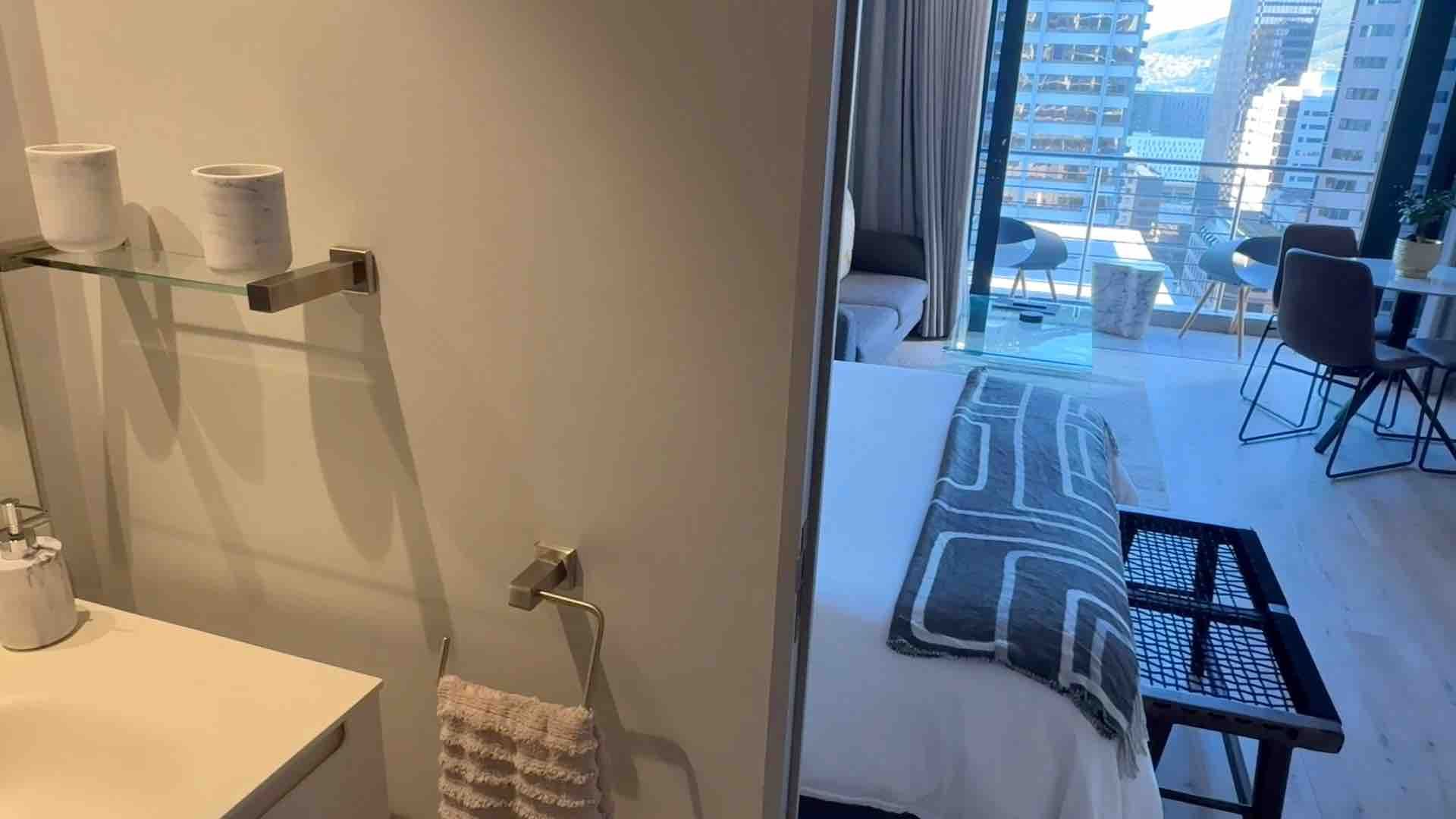 Bathroom with glass shower and towel beside a bedroom, showing a walkway to a city-view window.