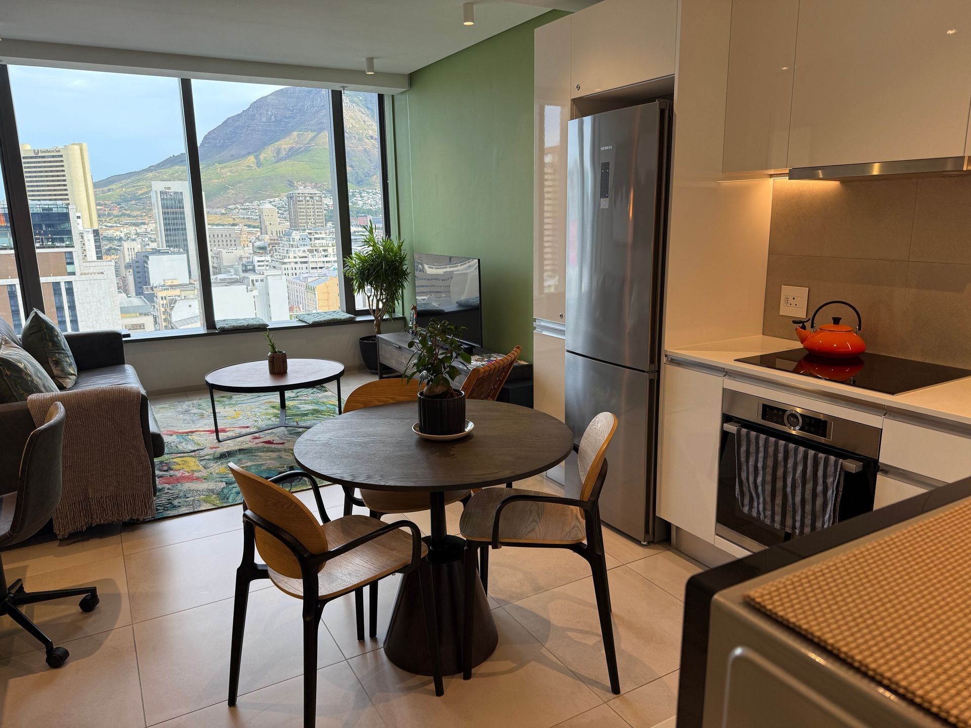 Modern apartment dining area with round table, kitchen, and large window view of the city and mountains