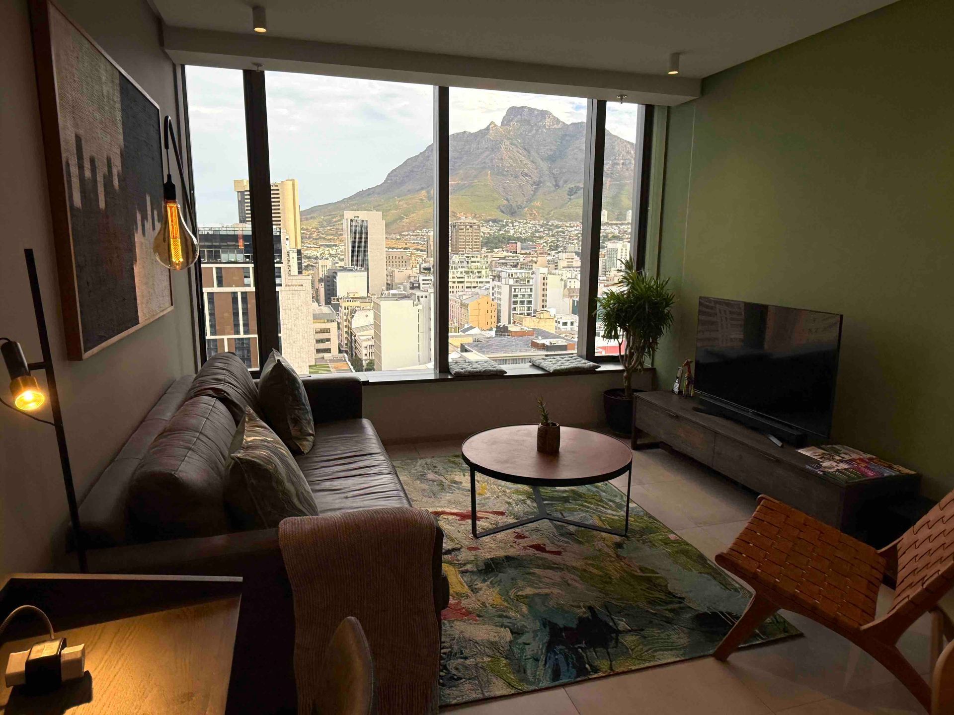 Modern living room with sofa, TV, coffee table, and large windows overlooking a city and mountains.