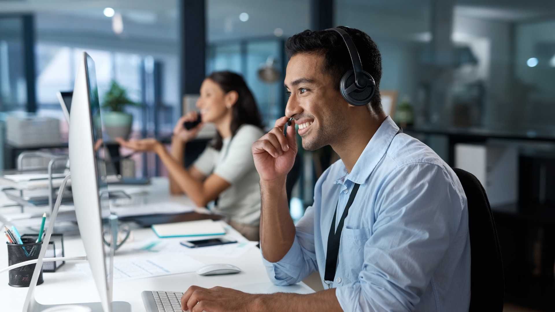 A man wearing headphones is talking on a cell phone while sitting in front of a computer.