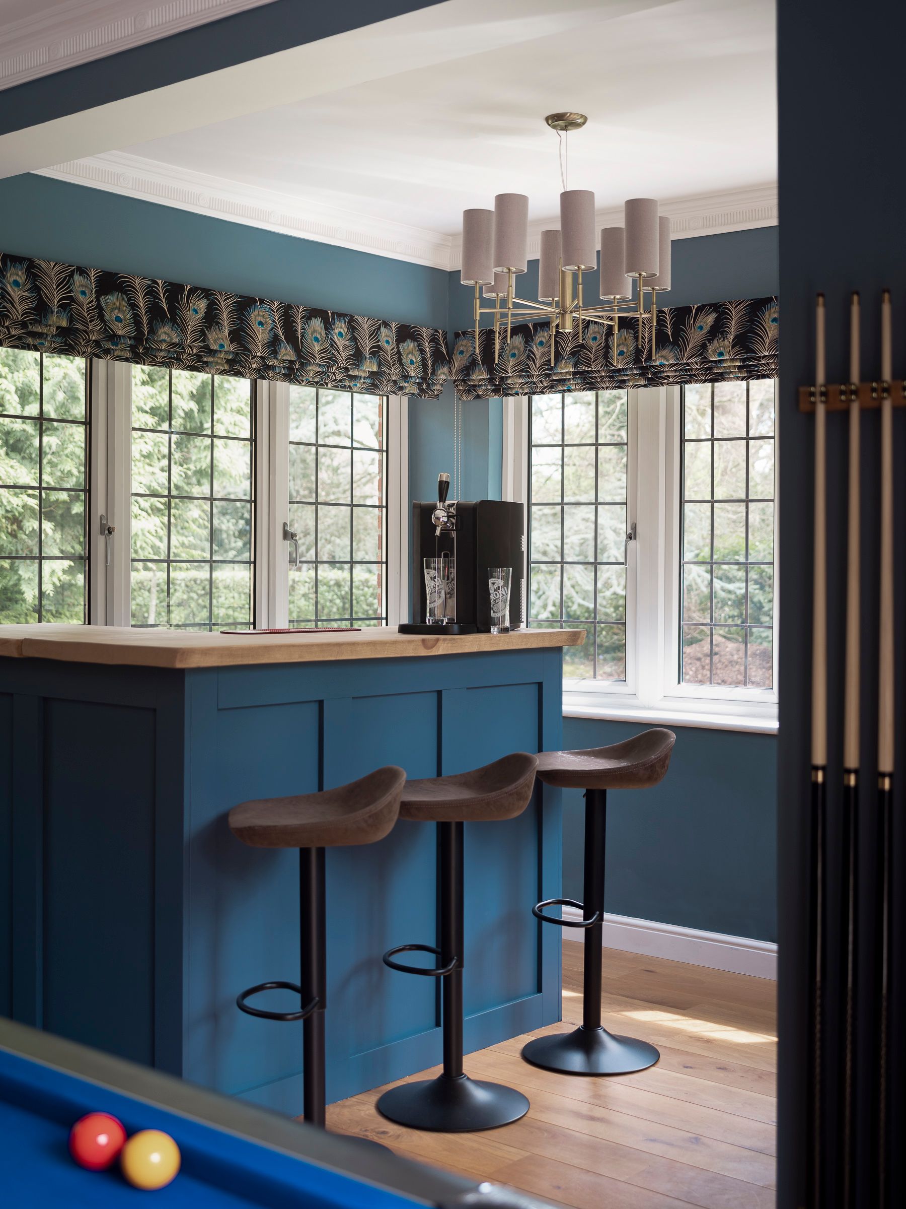 Bespoke blue games room bar with timber worktop, leather bar stools and peacock‑print pelmet overlooking the garden.