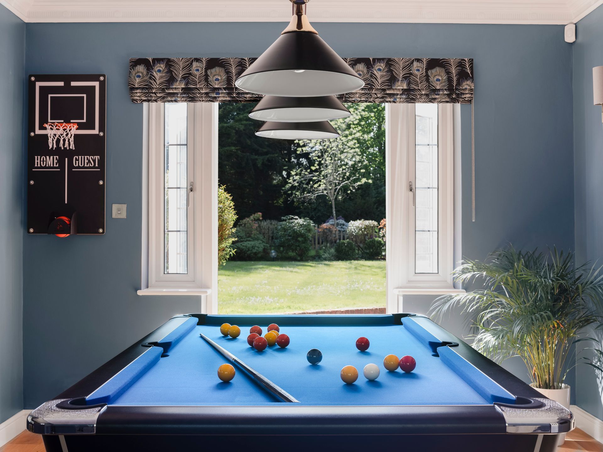 Pool table with blue cloth facing garden view, basketball hoop scoreboard and peacock‑print pelmets in blue games room.