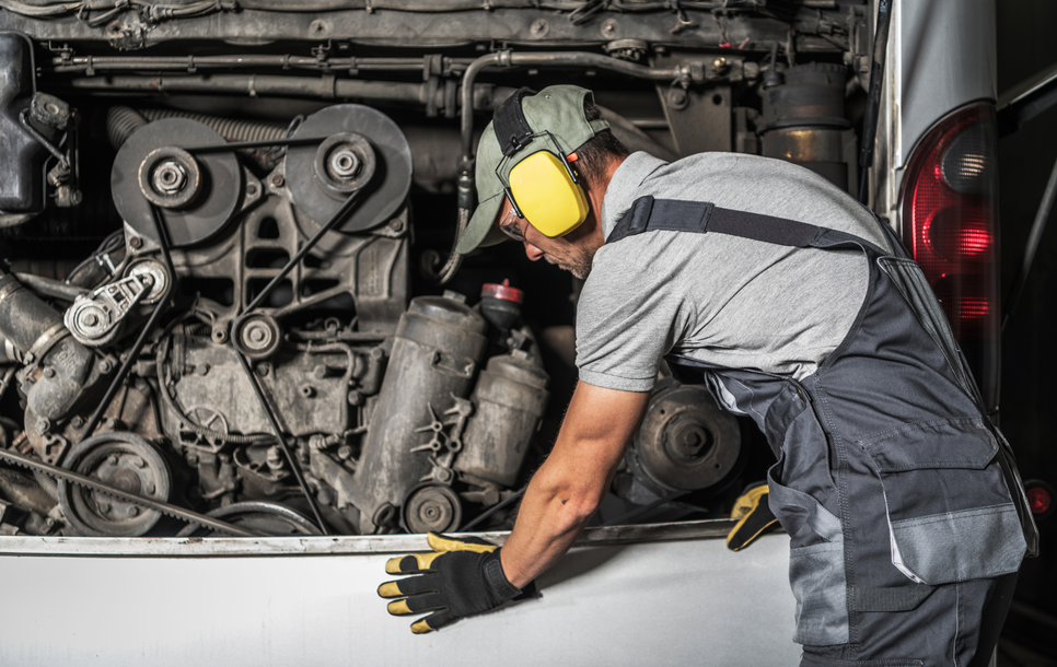 Mechanic working on a vehicle engine in a repair shop, wearing ear protection and gloves