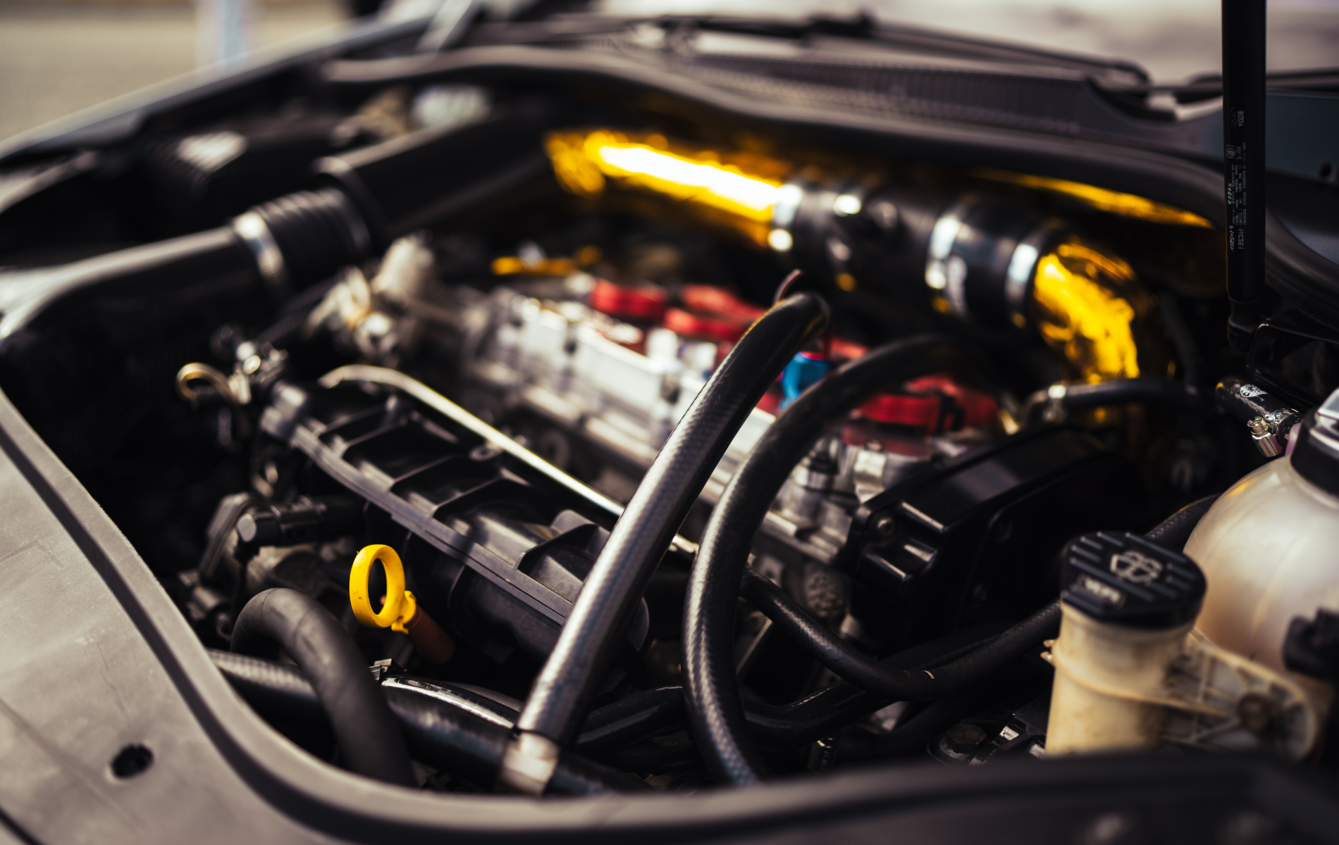 Car engine bay with exposed hoses, engine components, and yellow accents under the hood