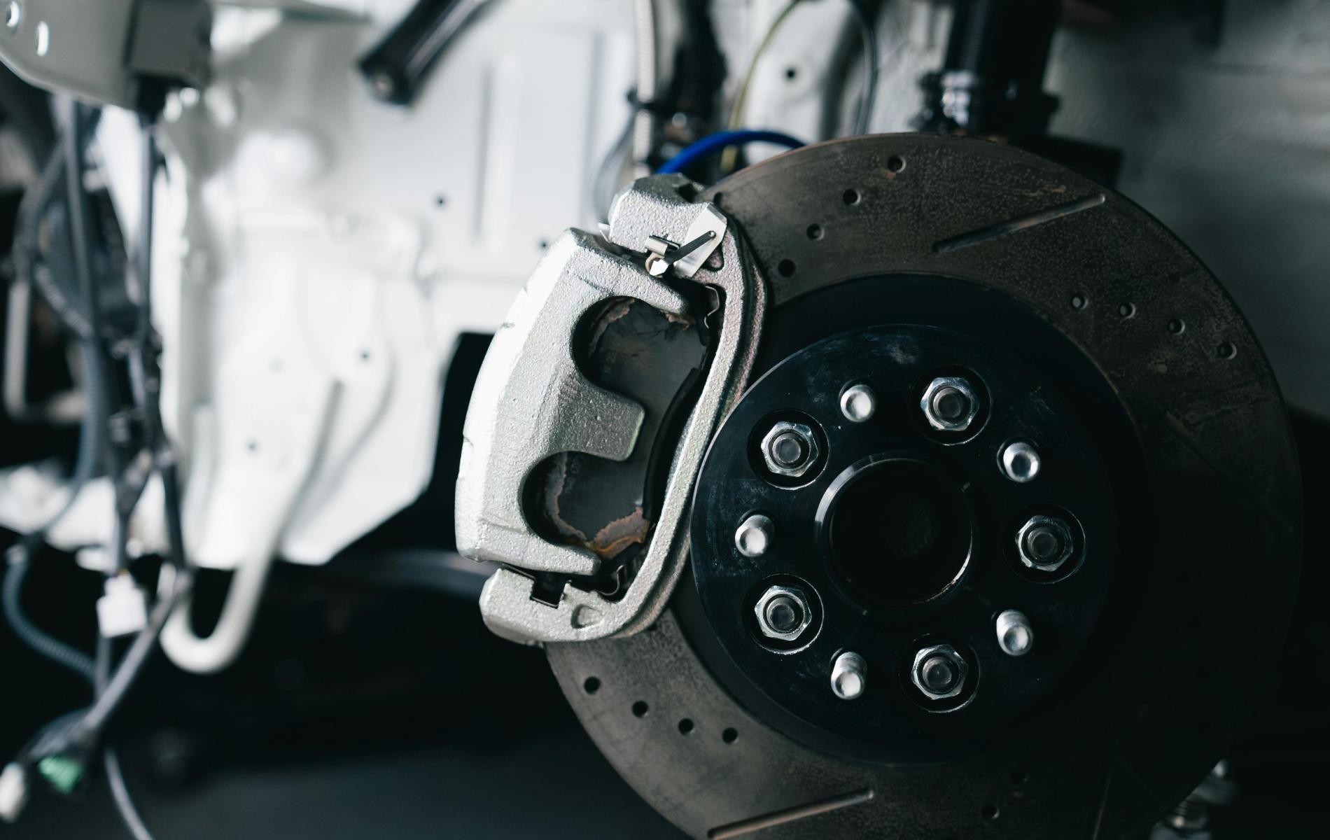 Close-up of a car brake rotor and caliper in a garage, with a white vehicle in the background