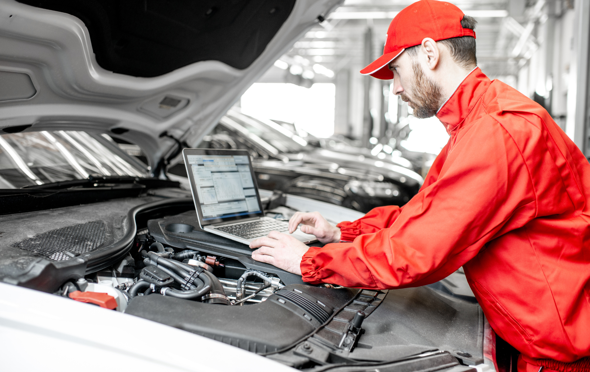 Mechanic in red uniform working under a car hood with a laptop in a garage