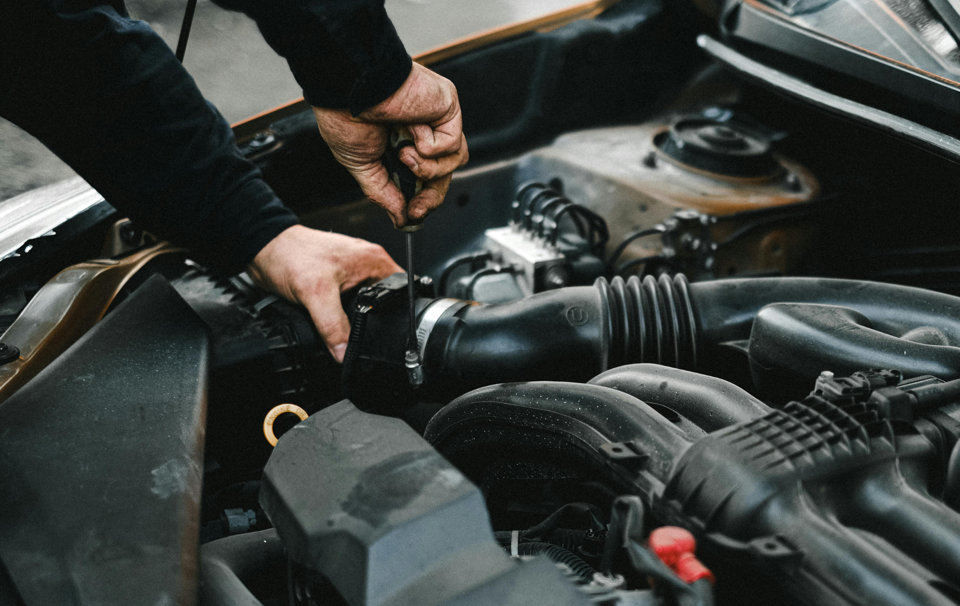 Hands working under a car hood, checking engine components near hoses and wiring.