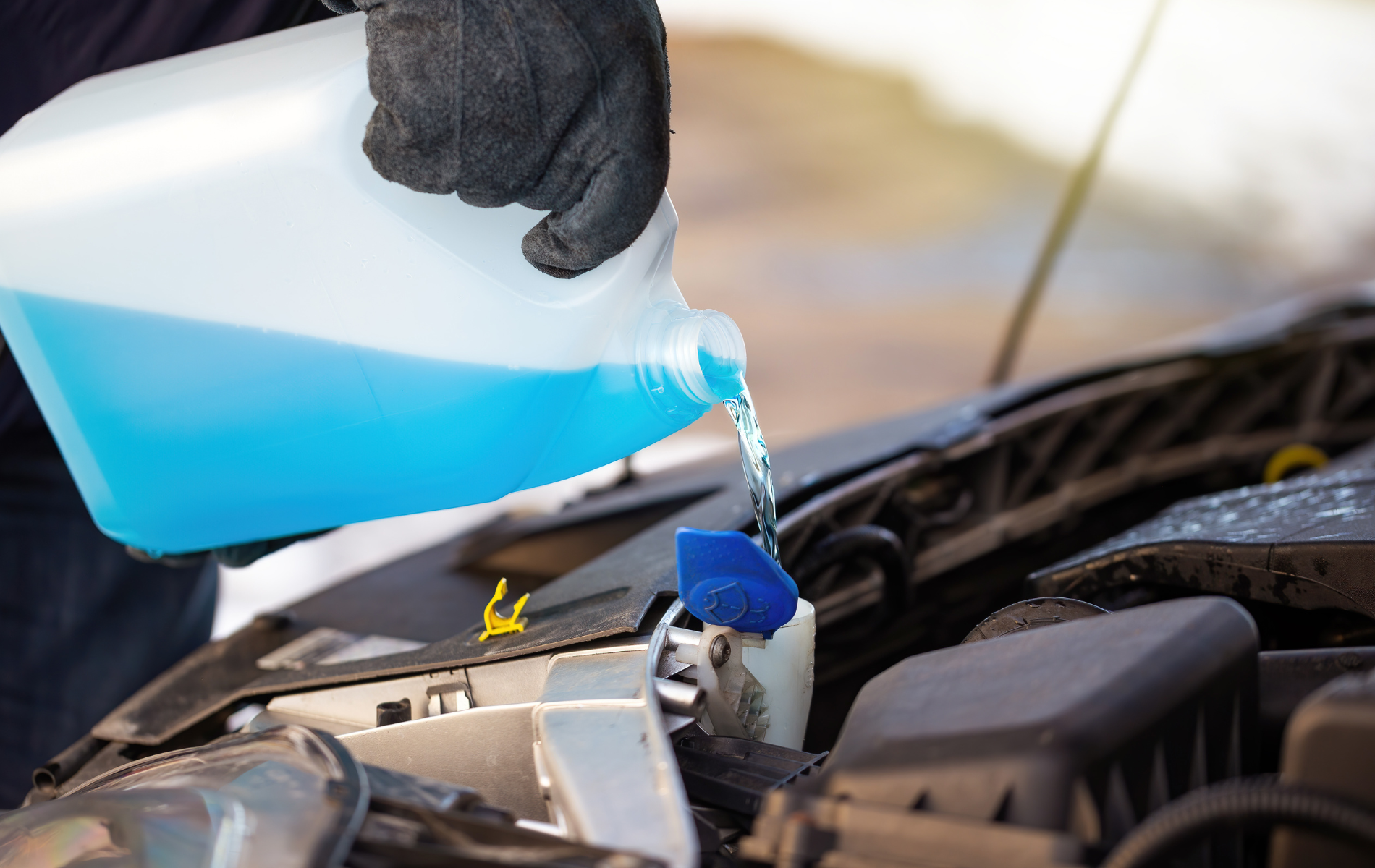Gloved hand pouring blue windshield washer fluid into a car engine reservoir