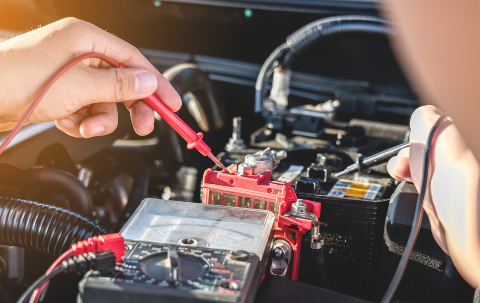Hands using a red multimeter to test a car battery in an engine bay