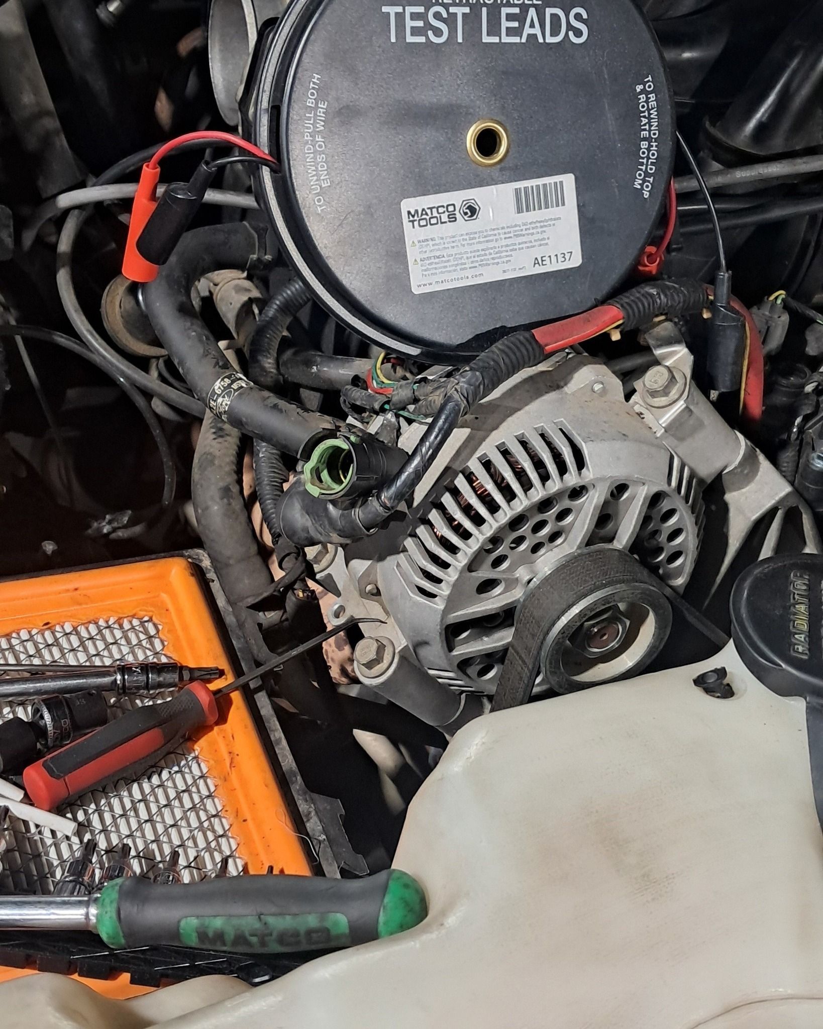 Close-up of a car engine bay with an alternator, test leads, and a coolant reservoir.
