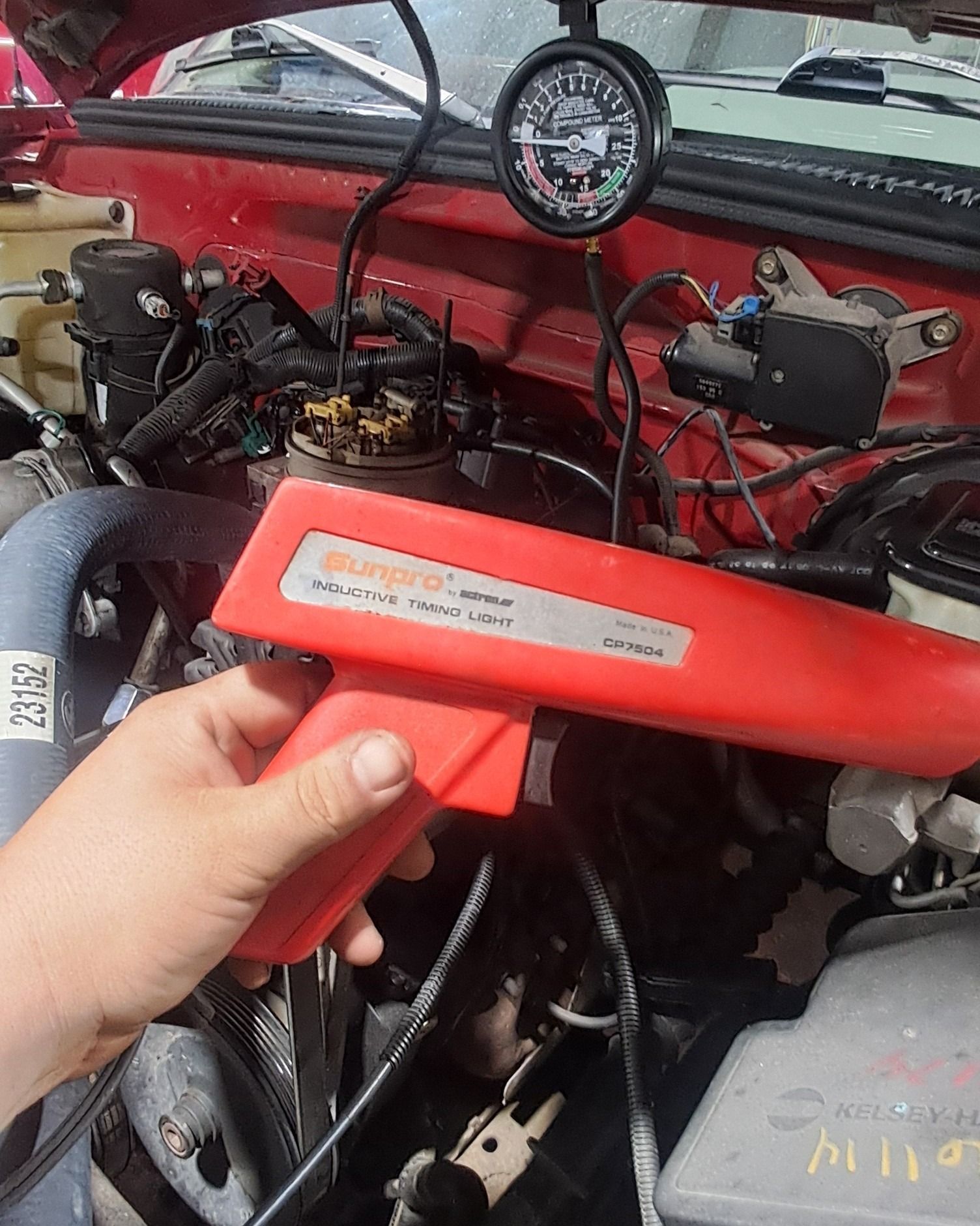 Hand holding a red timing light under a car hood, with engine bay and gauge visible