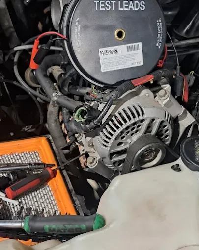 Car engine bay with alternator, test leads, and an orange tool tray beside a coolant reservoir