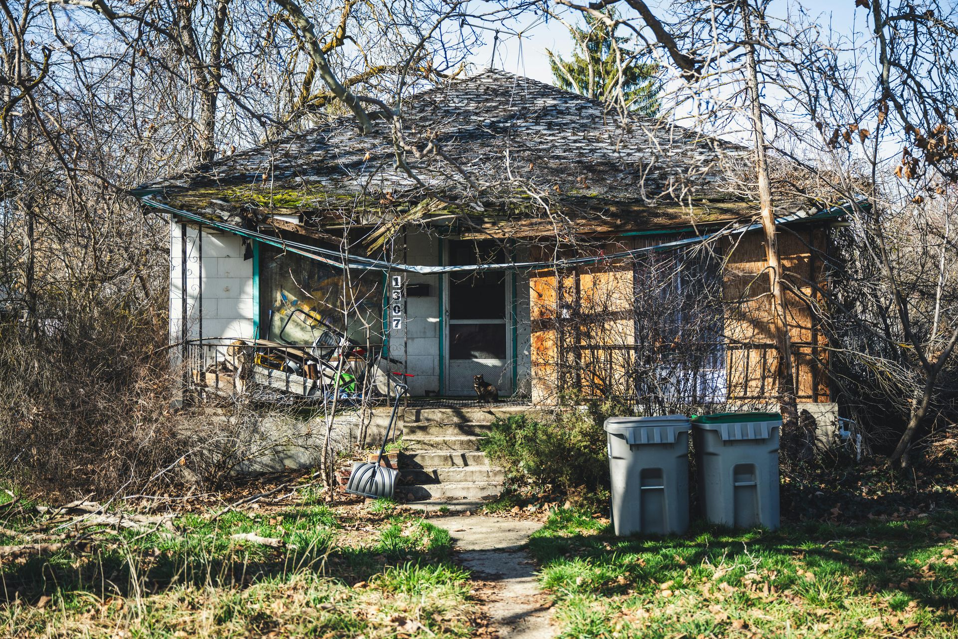 A small, weathered house with an overgrown yard and two gray trash bins in the foreground.