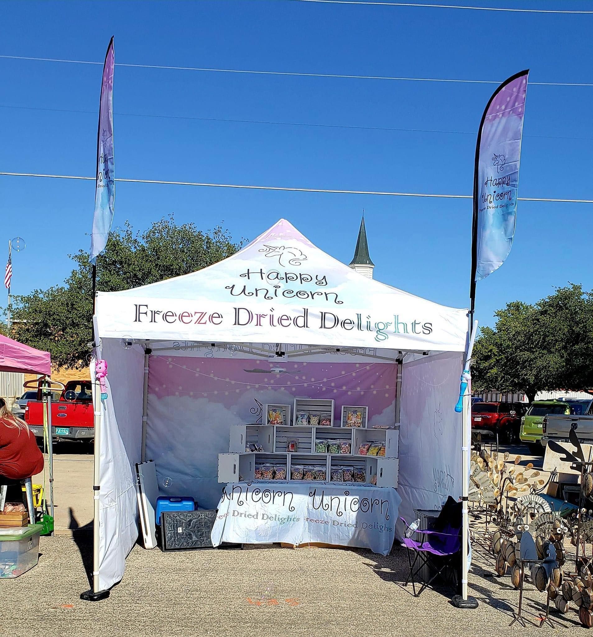 Outdoor market booth with a white canopy and purple flags, displaying items on a table under a bright blue sky