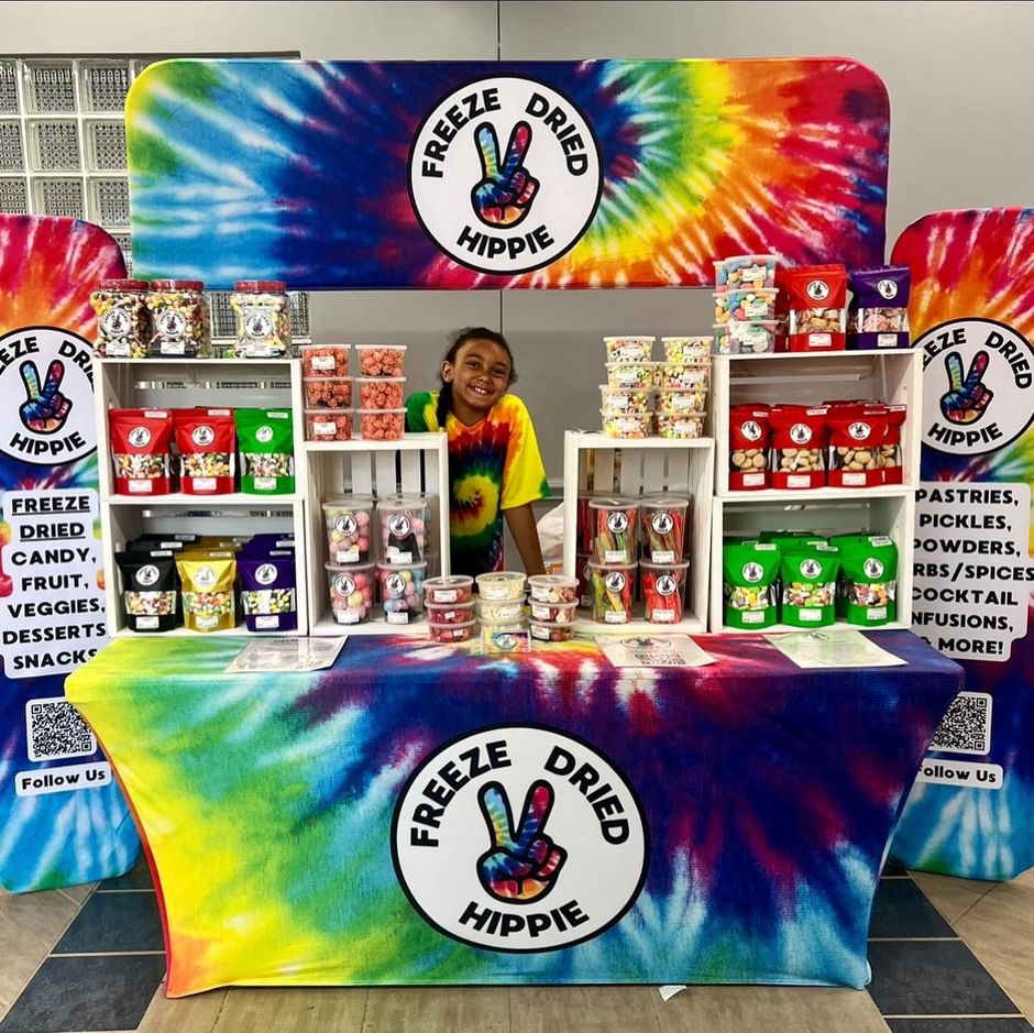 Vendor booth with rainbow tie-dye “Freeze Dried Hippie” display and shelves of colorful snack packages.