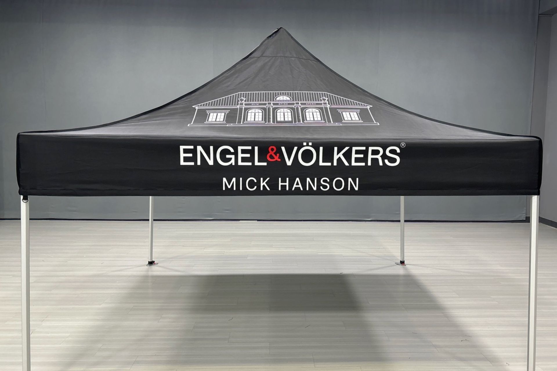 Black Engel & Völkers pop-up canopy tent with “Mick Hanson” text in a gray indoor space