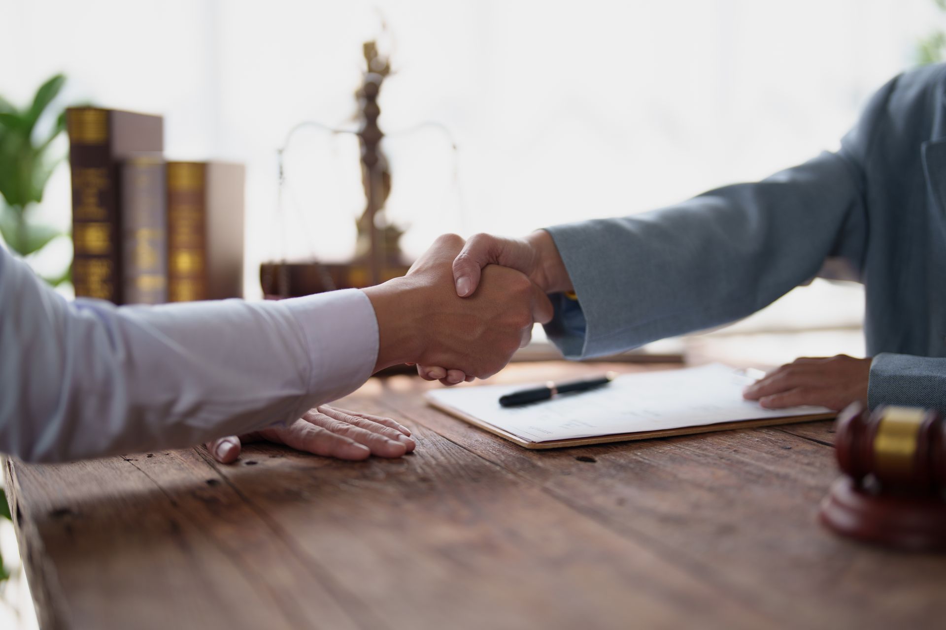 Two people shake hands across a wooden desk with a pen and papers in a bright office.