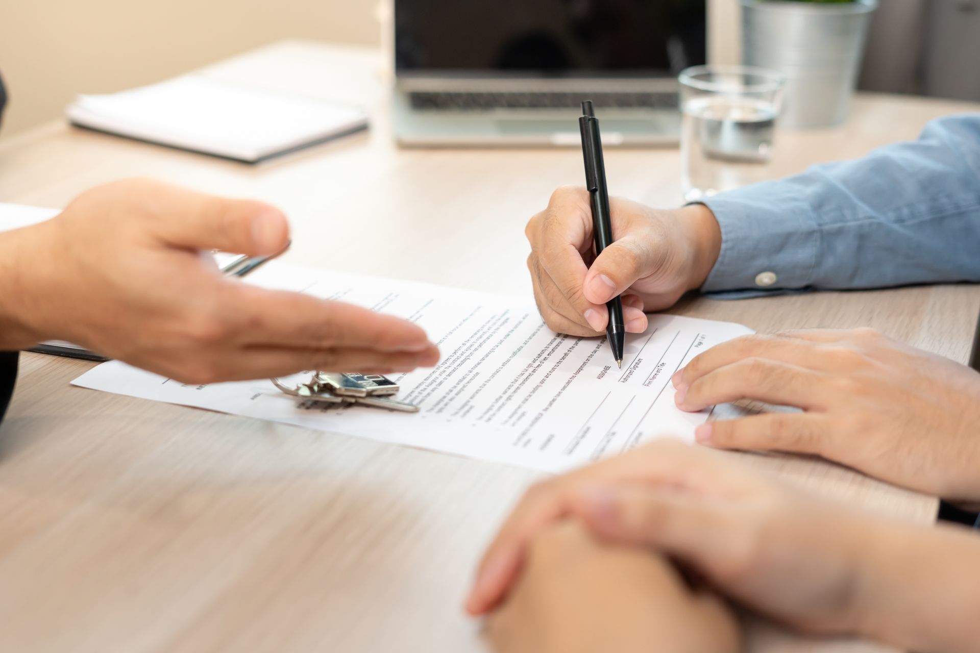 Hands signing a document across a desk with keys, papers, and a glass in the background Hands signing a document across a desk with keys, papers, and a glass in the background