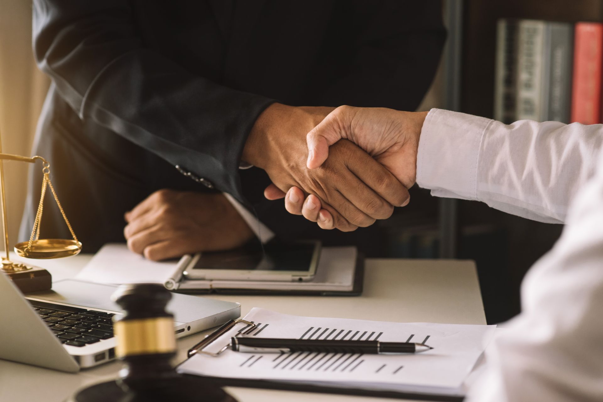 Businesspeople shaking hands over a desk with contract papers and a laptop Businesspeople shaking hands over a desk with contract papers and a laptop