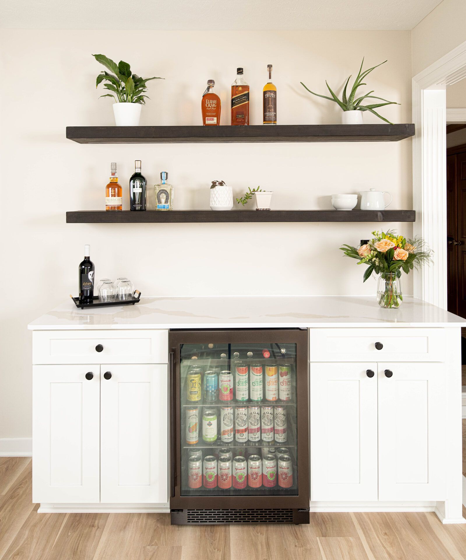 Home bar with white cabinets, built-in beverage cooler, wood shelves displaying liquor and plants.