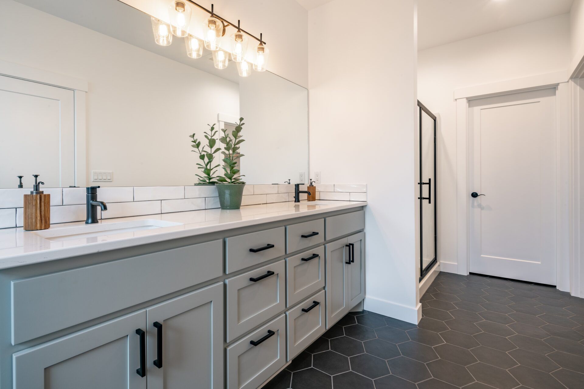 Modern bathroom with gray vanity, white countertops, black hexagon floor tiles, and a glass shower.
