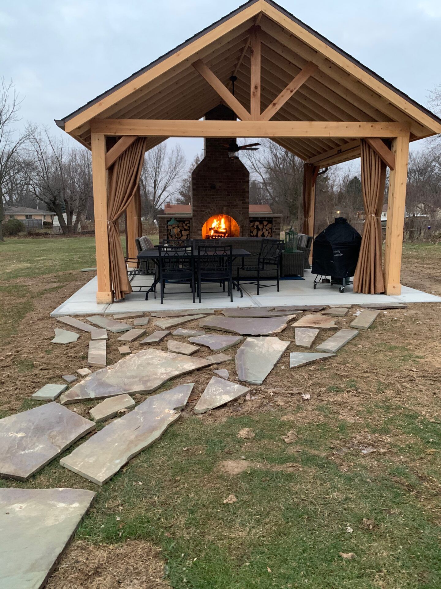 Outdoor pavilion with fireplace, seating, and flagstone path. Wooden structure with curtains, grill, and fire burning.