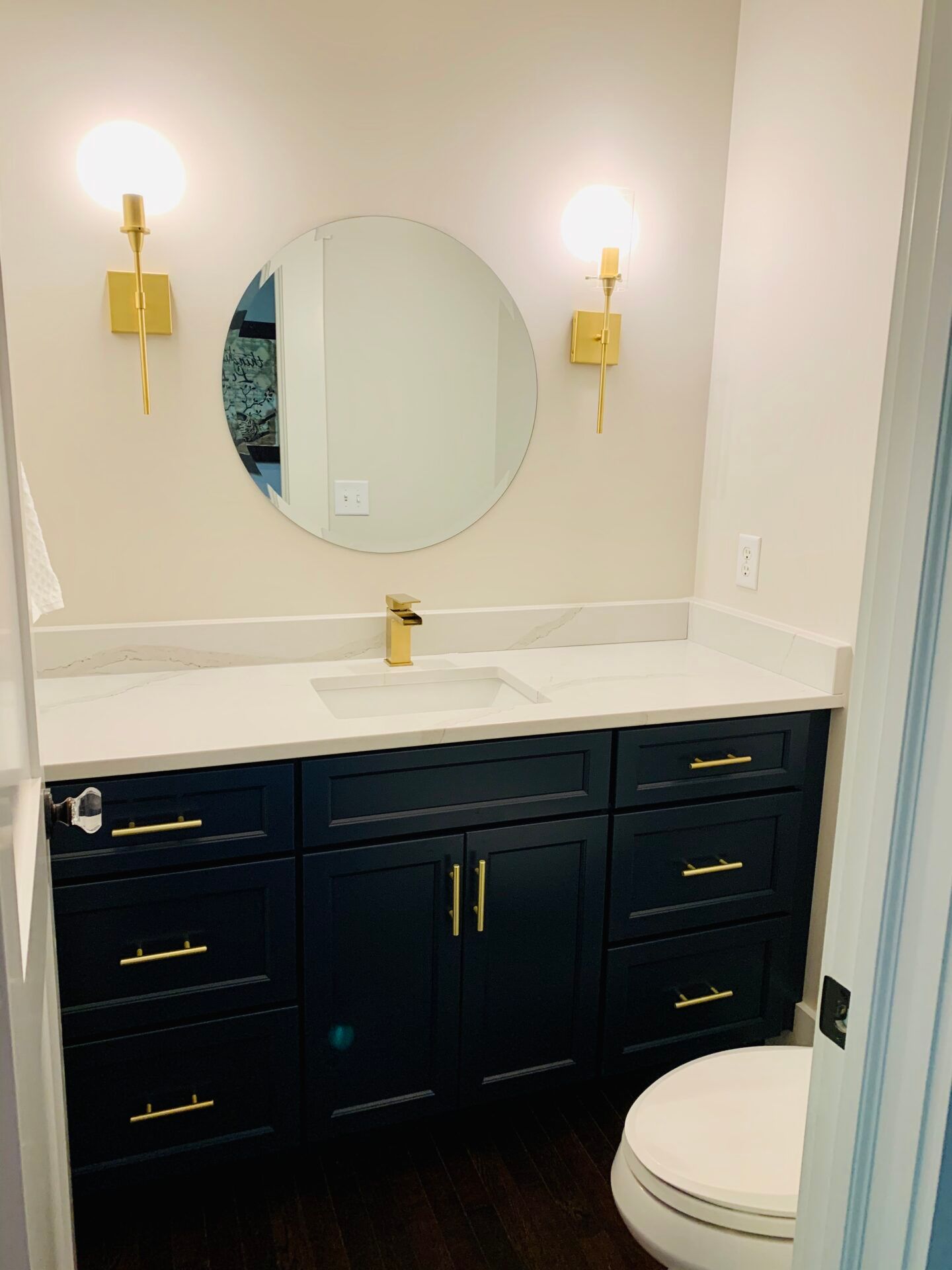 Bathroom with dark blue vanity, gold fixtures, white countertop, round mirror, and toilet.