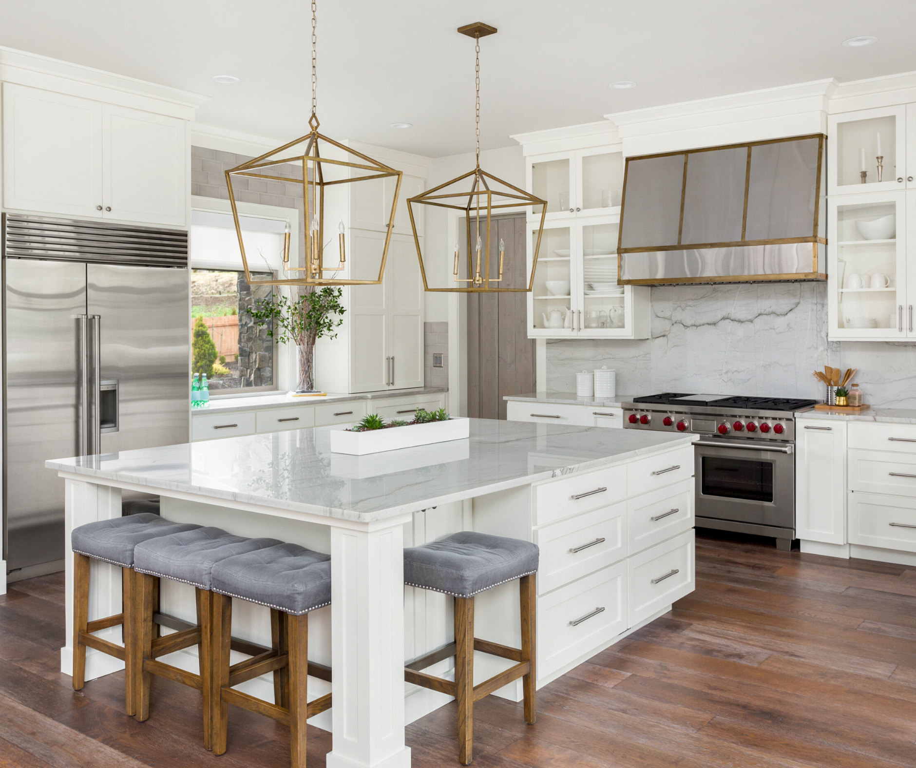 Luxurious white kitchen with island, marble countertops, pendant lights, stainless steel appliances, and wood floors.
