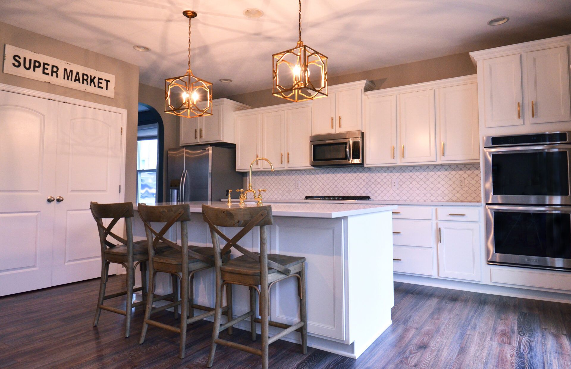 A bright kitchen with white cabinets, island, and gold light fixtures. Wooden chairs sit at the island.