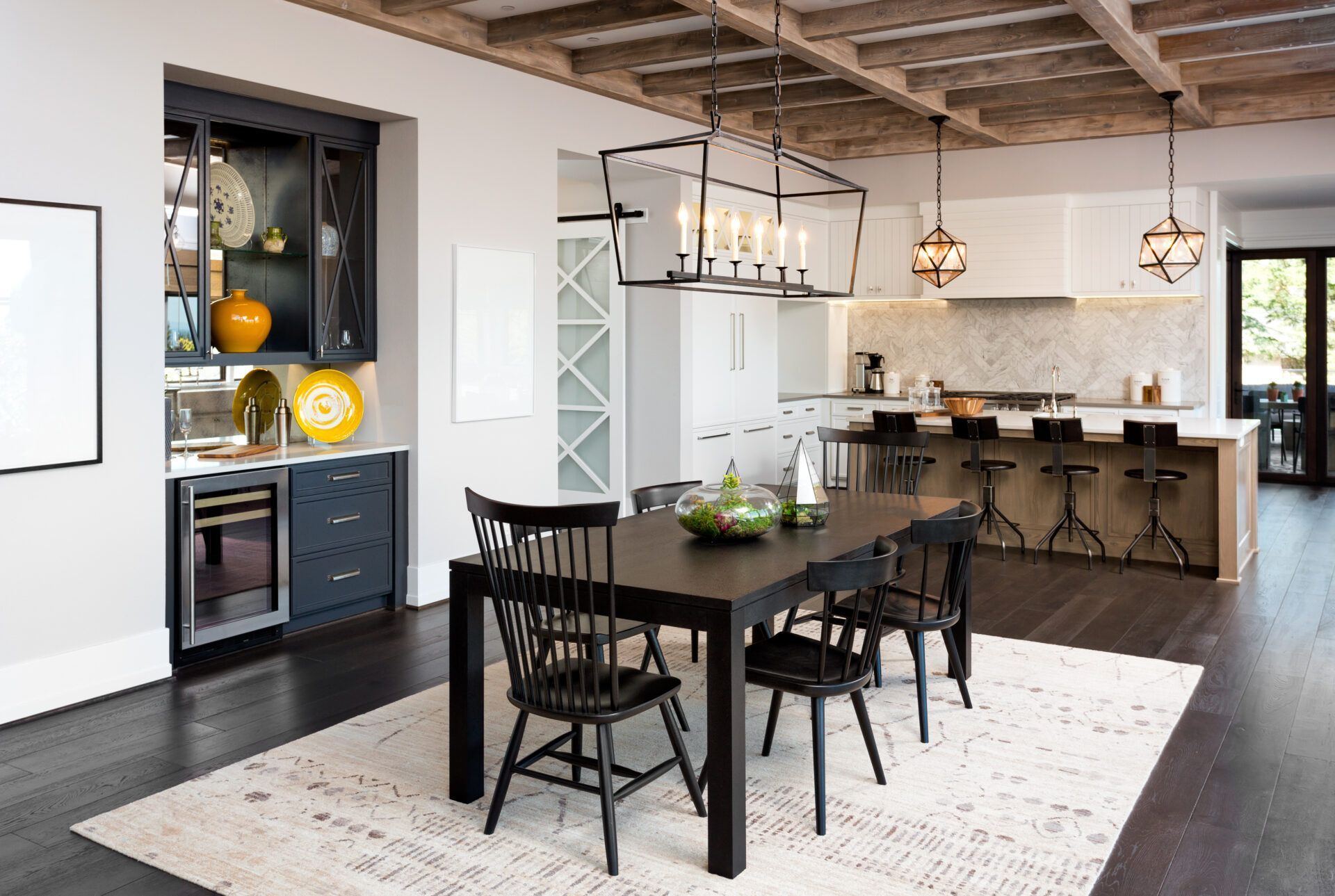 Dining room with black table and chairs, kitchen in the background, hardwood floors, and exposed beam ceiling.