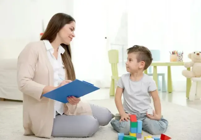 Woman with clipboard talks to boy playing with blocks in a playroom.