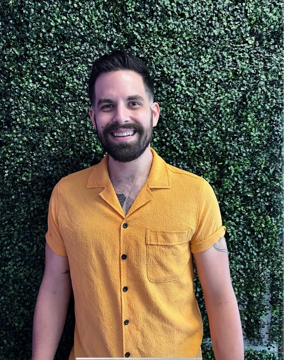 Smiling bearded man in a mustard shirt standing in front of a green hedge wall