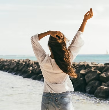 Woman with arms raised, standing on a rocky beach, facing the ocean.