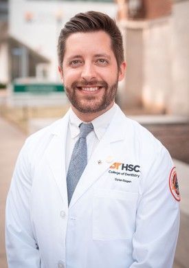 A professional headshot of a smiling person wearing a white lab coat with an HSC College of Dentistry logo and a tie.