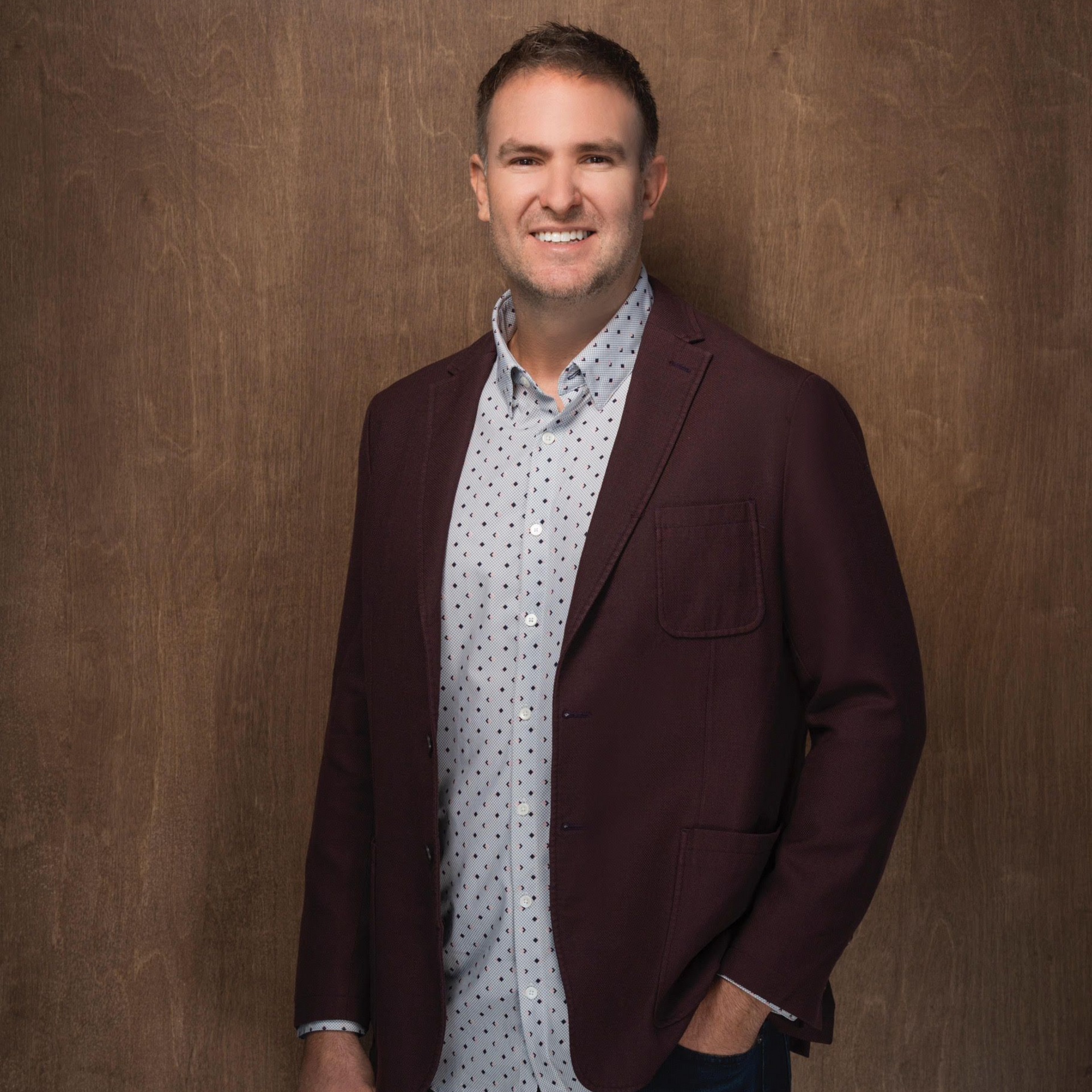 A smiling person in a dark red blazer, patterned button-down shirt, and jeans, standing against a brown textured backdrop.