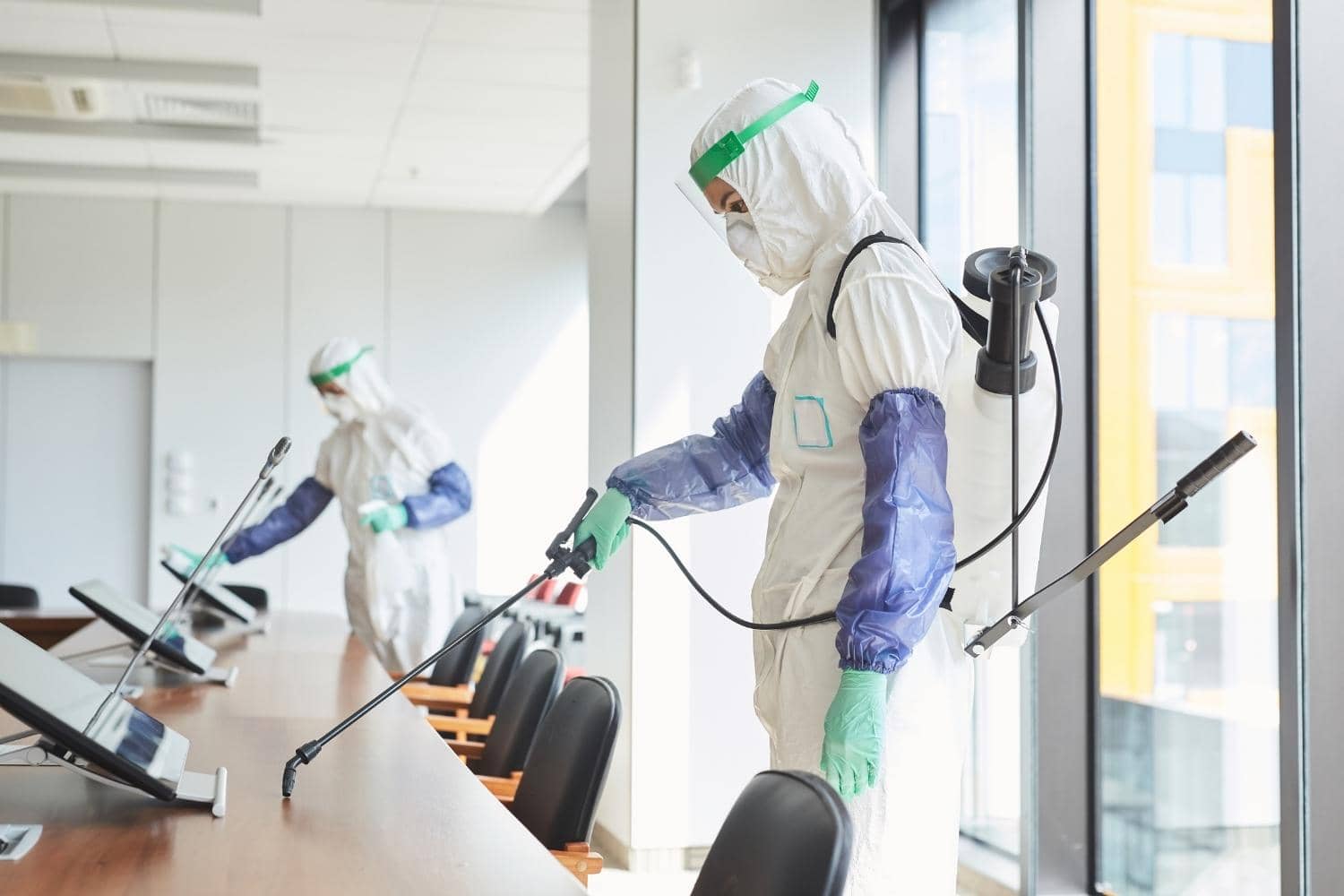 Two people in protective suits are disinfecting a conference room.