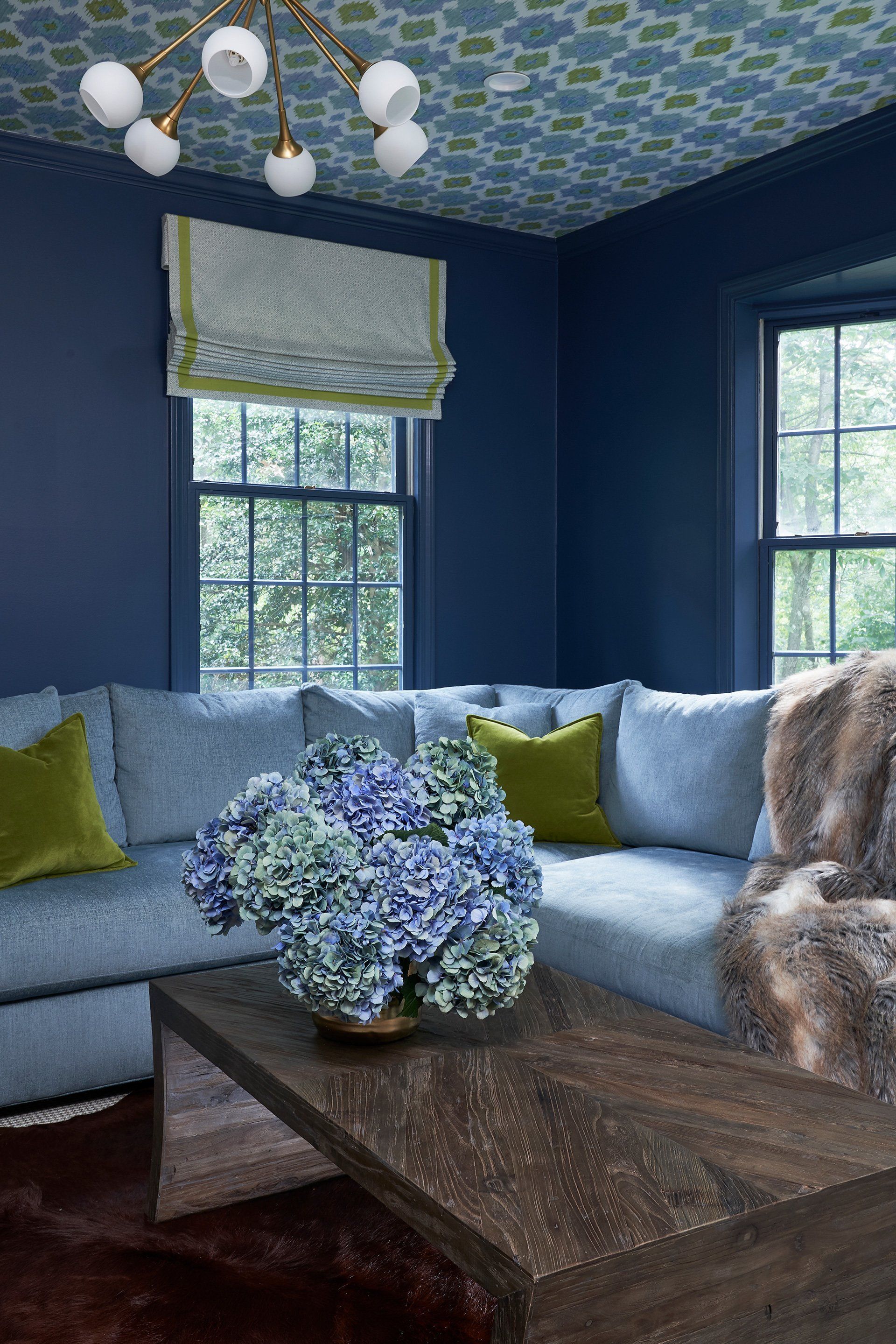 Blue-walled room with floral ceiling, blue sectional sofa, hydrangea centerpiece, and wooden coffee table.