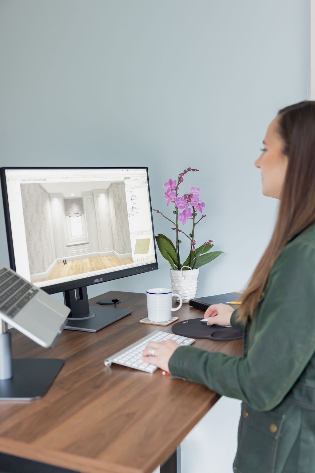Woman working at standing desk with computer, a laptop, and a purple orchid.