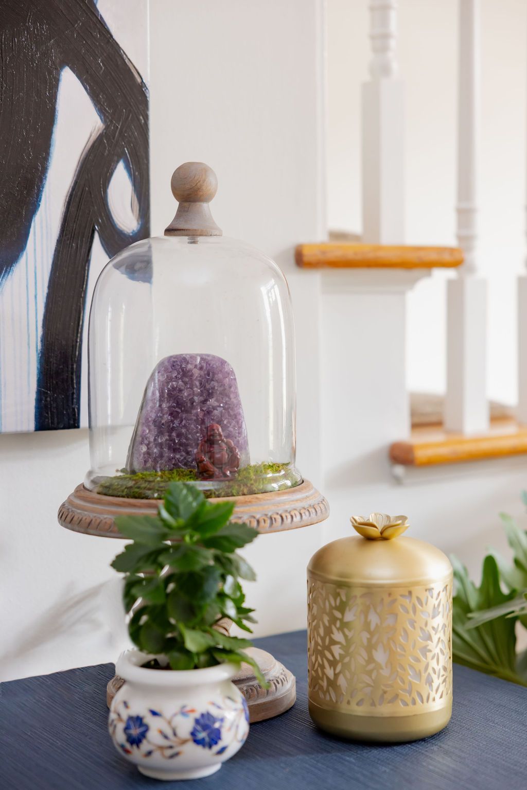Amethyst geode in glass cloche on a table next to a potted plant and gold candle holder.