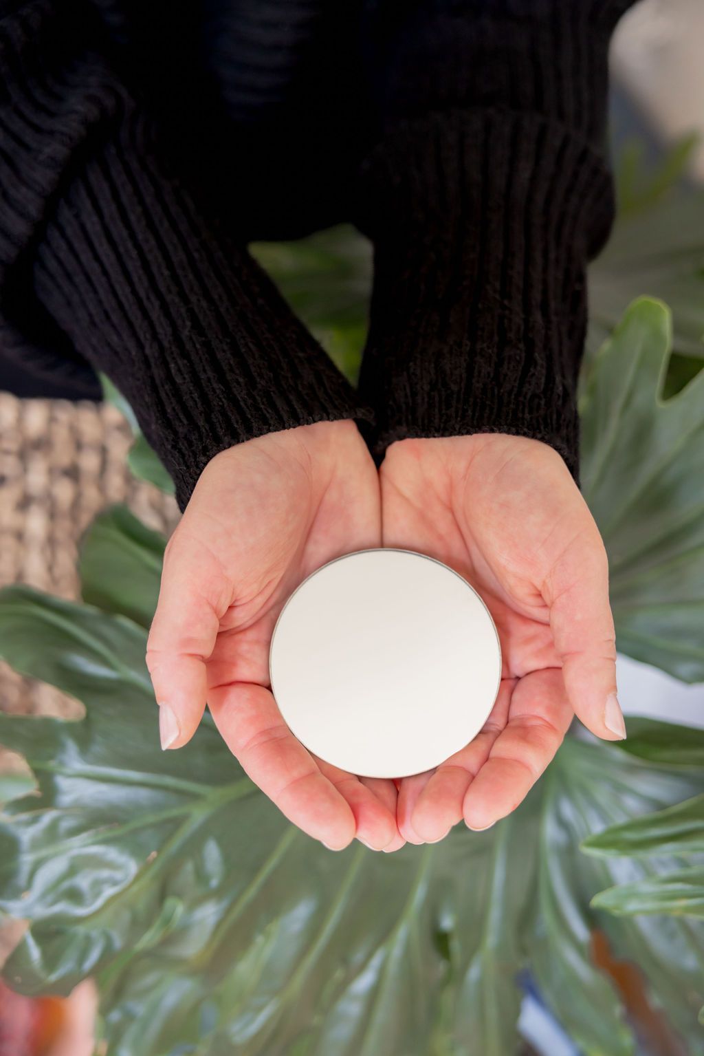 Hands holding a white circular object, possibly a cosmetic product, against a backdrop of green leaves.