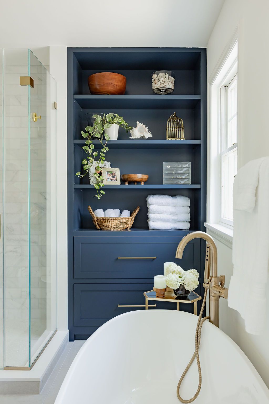 Blue built-in bathroom shelving with décor, gold faucet, and white tub. Next to shower with glass door.