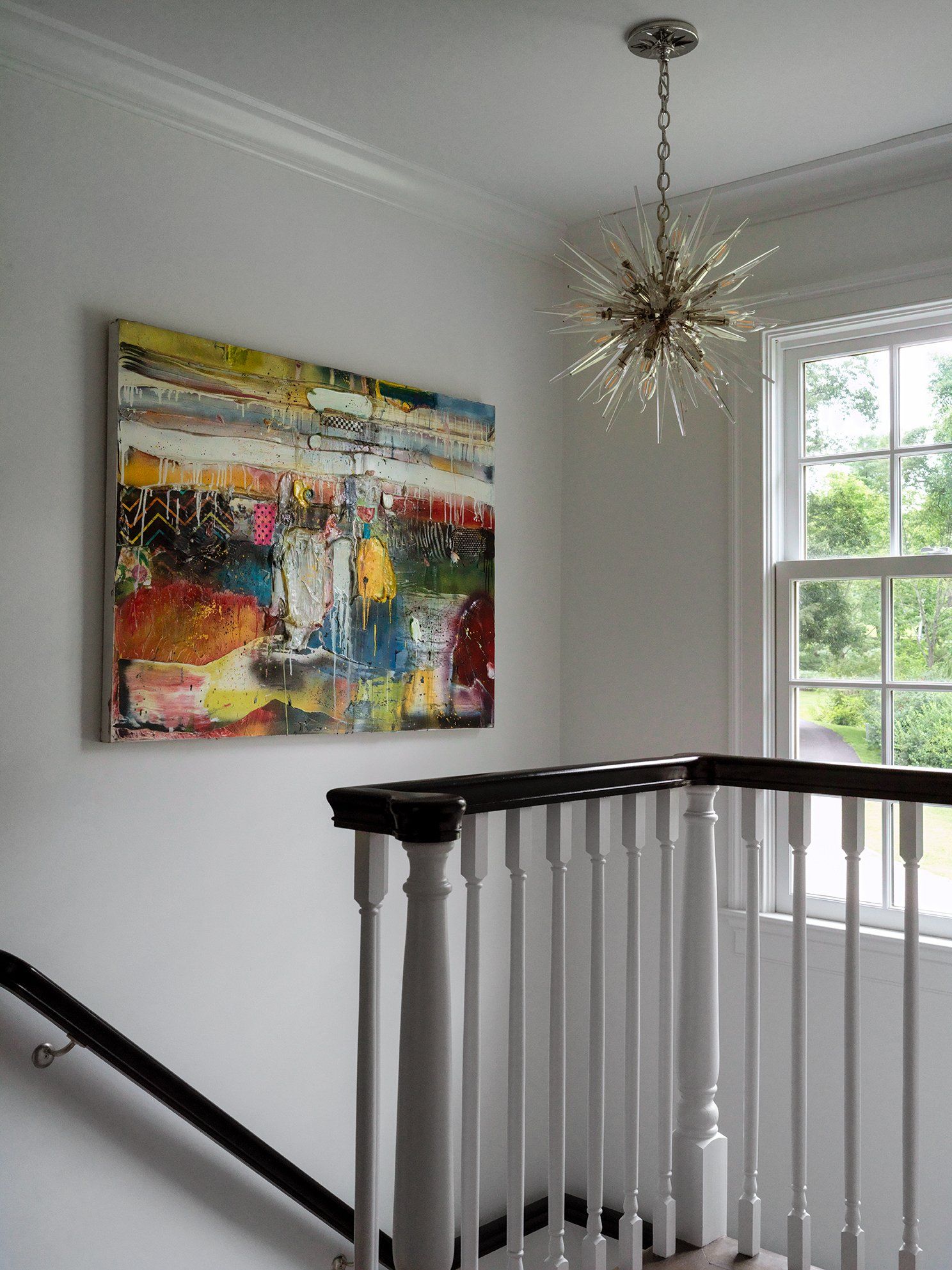 Abstract art on a white wall above a staircase with a modern chandelier and window.