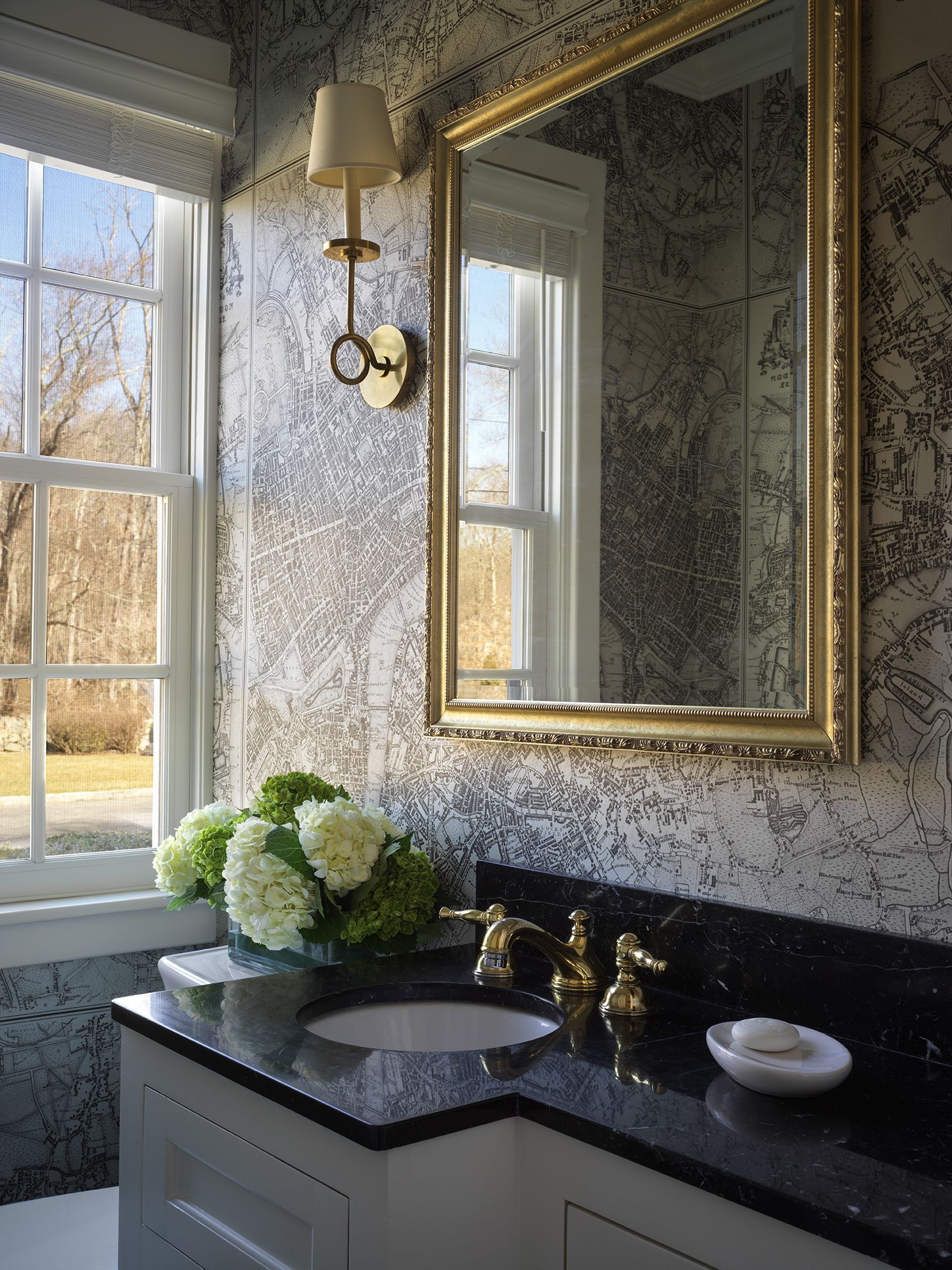 Bathroom with patterned wallpaper, gold-framed mirror, white cabinetry, black countertop, and floral arrangement.
