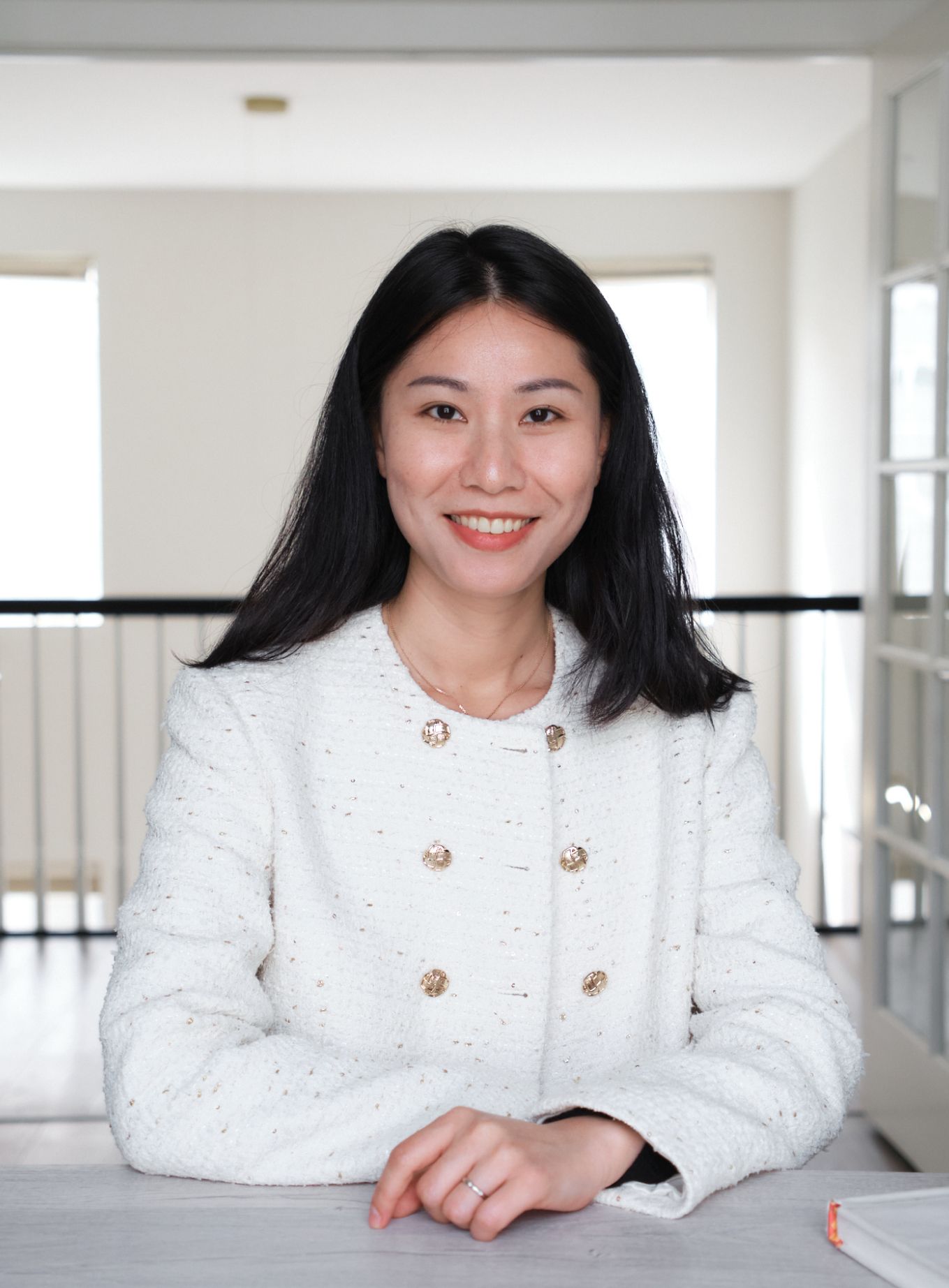A woman in a white jacket is sitting at a table with her hands folded.