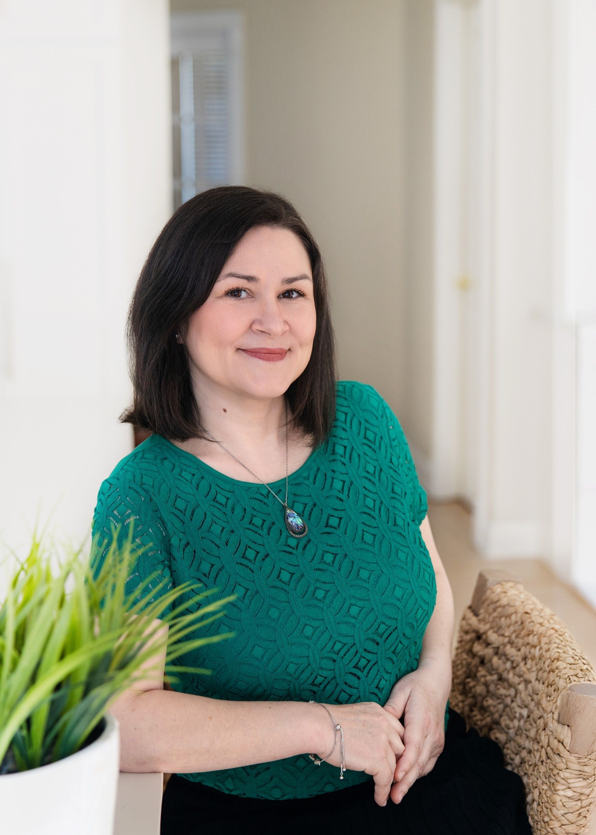 Woman seated in a bright hallway, wearing a green top and smiling beside a potted plant.