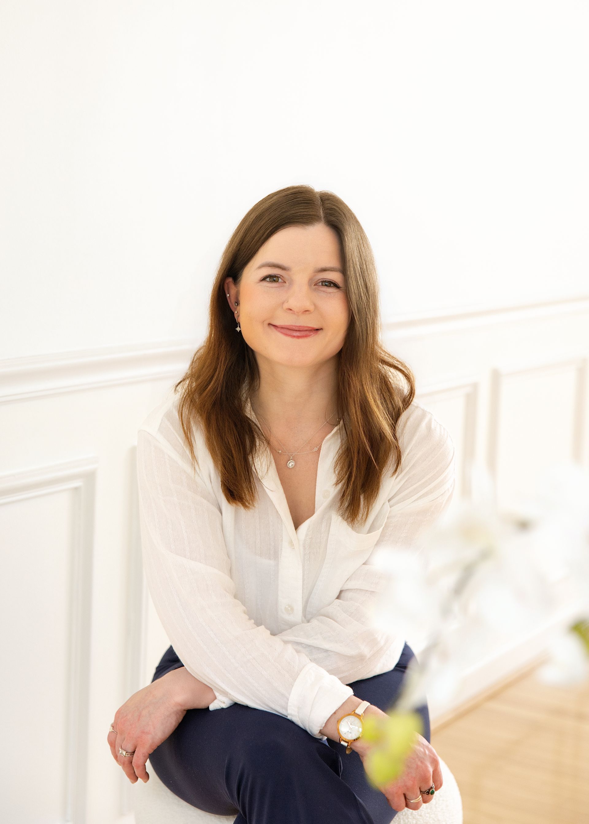 Smiling woman seated indoors against a white wall, wearing a white blouse and dark pants.