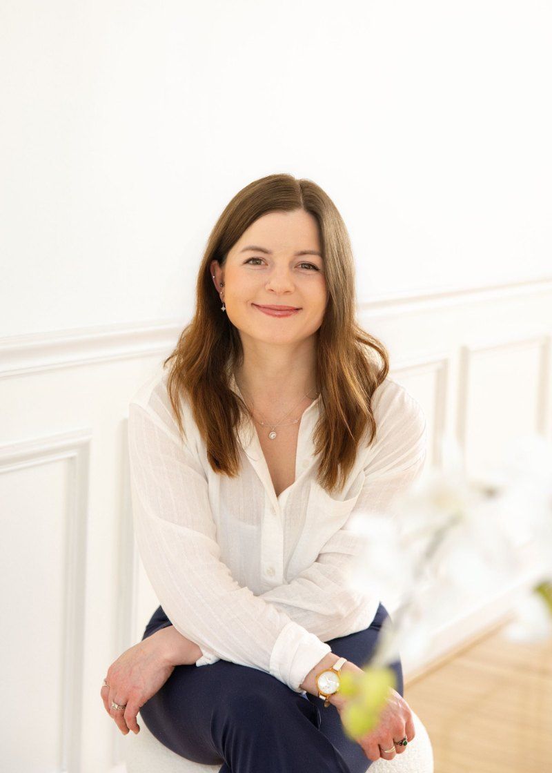Smiling woman seated indoors against a white wall, wearing a white blouse and dark pants.